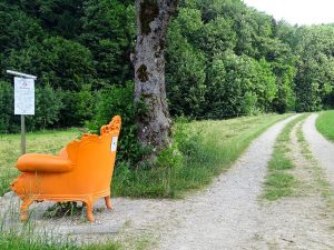 Auf der linken Seite des Bildes befindet sich ein ganz orange bemalener Sessel. Dahinter geht eine Schotterstraße vorbei und ein Baum steht daneben.