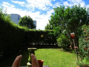 Der Blick von meiner Terrasse auf den Minigarten. Im Vordergrund Deko-Material und rotes Herz am Rosenstock. Im Hintergrund das Nachbarhaus und blauer Himmel mit Schäfchenwolken.