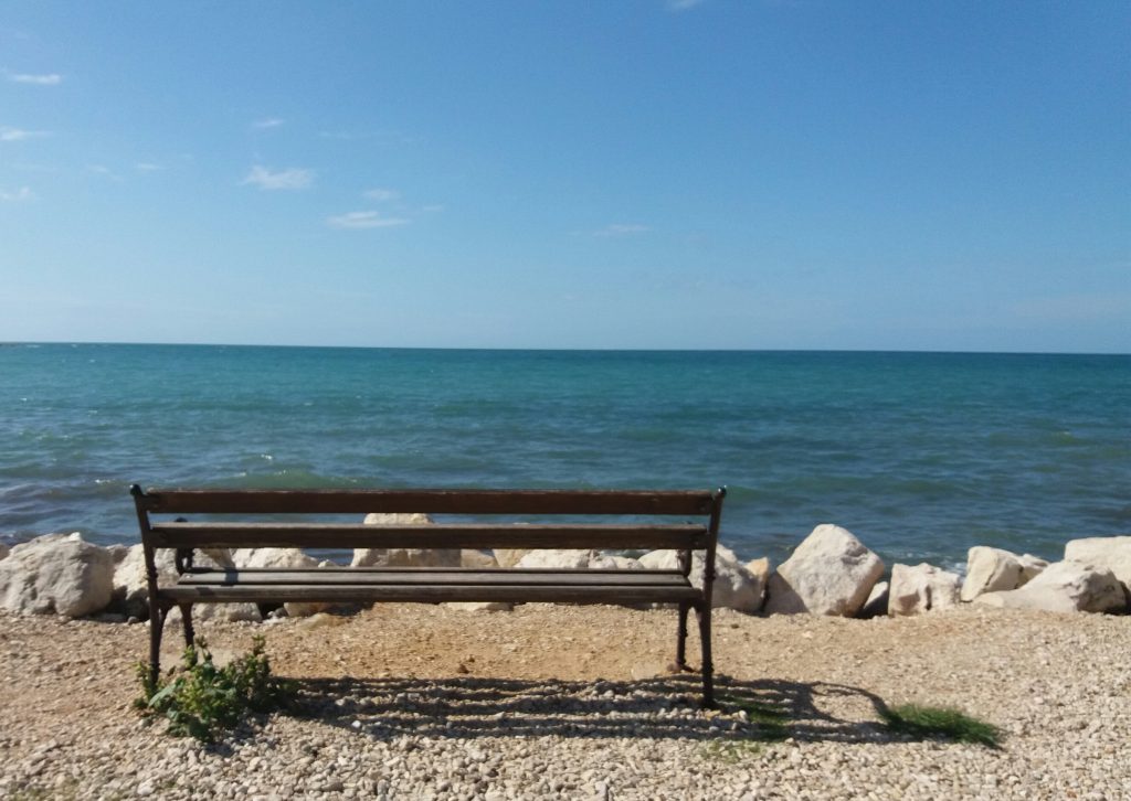 Ein Bankerl von hinten fotografiert mit dem Blick aufs Meer und den blauen Himmel