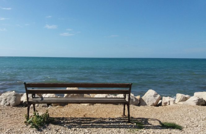 Ein Bankerl von hinten fotografiert mit dem Blick aufs Meer und den blauen Himmel