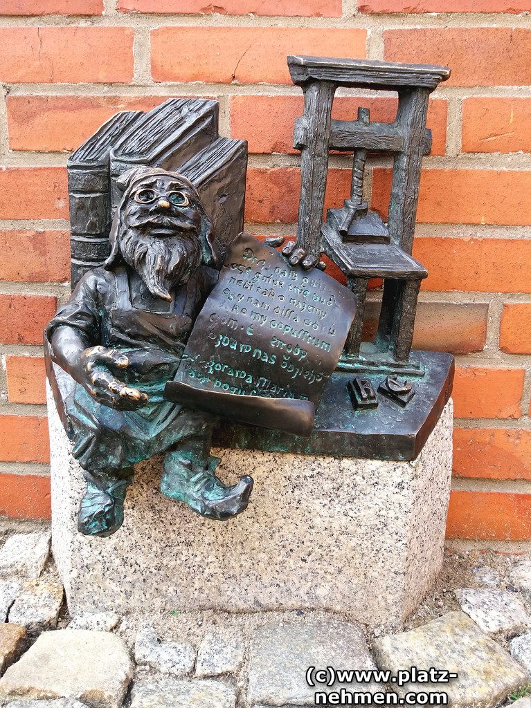 Der Buchdrucker Zwerg sitzt auf einem Sockel und lehnt an einigen Büchern. In der Hand hat er ein Papier mit Text. Im Hintergrund die Buchpresse.