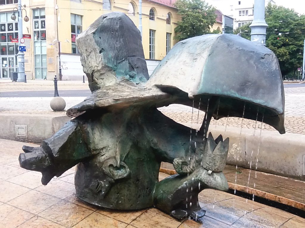 Ein Zwerg mit riesigem Hut hält seinen Schirm über den Froschkönig in einem Brunnen