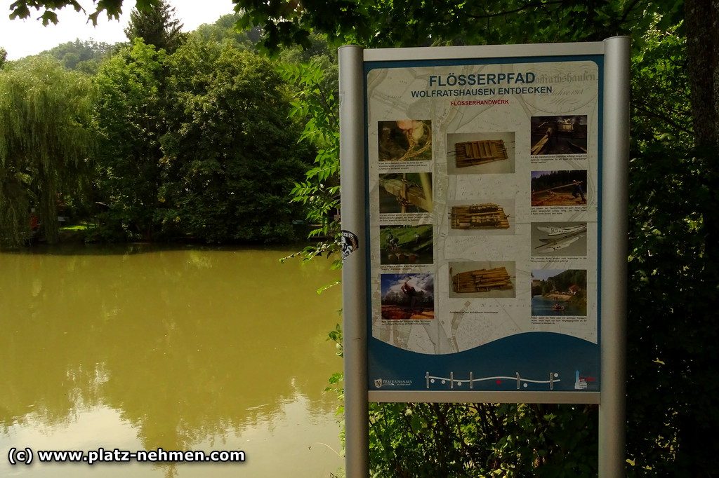 Der Fluß Loisach im Hintergrund mit Bäumen und im Vordergrund ein Bild, dass über den Flößerpfad informiert